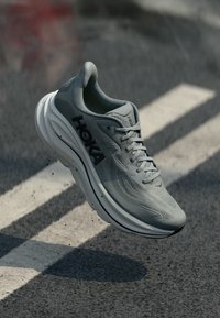 Sapatilha de corrida Hoka cinza claro com sola branca espessa, atacadores atados, pairando acima de uma estrada de asfalto com linhas brancas e sombras.