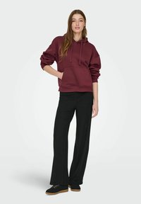 Bordeauxrode capuchontrui met een zak aan de voorkant, gecombineerd met zwarte ribgebreide broek met wijde pijpen en zwarte schoenen met rubberen zolen.