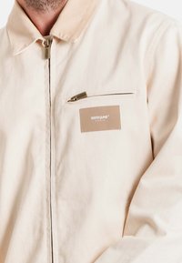 Beige jas van textuurstof, voorzien van een rits aan de voorkant, een borstzak met een rits en een merketiket in een contrasterende kleur.