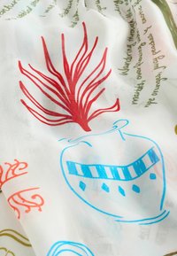 Biela tkanina s červeným dizajnom morských rias, modrý náčrt vázy a ručne písaný zelený text čiastočne viditeľný na povrchu.