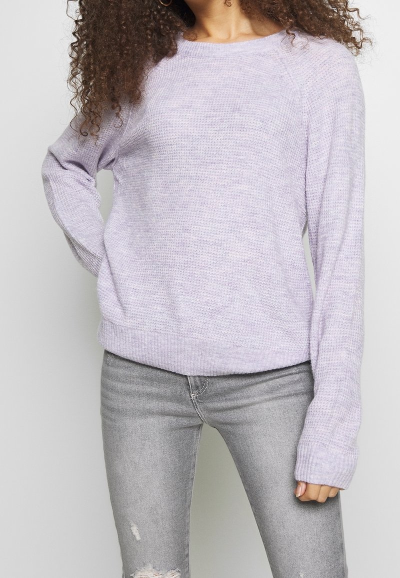 Femme aux cheveux bouclés portant un pull en maille violet clair et un jean gris déchiré, posant avec une main sur la hanche.