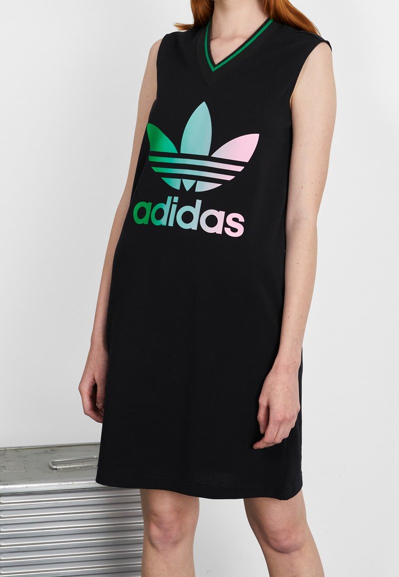 Femme portant une robe noire sans manches Adidas avec un grand logo dégradé pastel à l'avant, debout près d'une valise argentée.