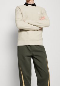 Jersey de punto beige claro con cuello redondo y textura acanalada, combinado con pantalones verde oliva que tienen franjas laterales marrón claro.