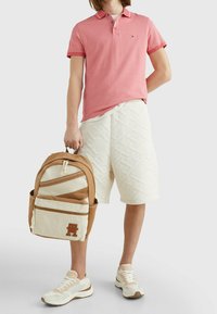 Mężczyzna w różowej koszulce polo, w fakturowanych białych szortach, w beżowo-białych sneakersach, trzymający beżowo-brązowy plecak za uchwyt.
