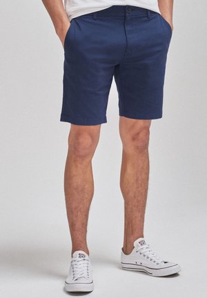 Homme portant un short bleu marine, des baskets blanches basses et une chemise blanche, les mains dans les poches, debout sur un fond gris clair.