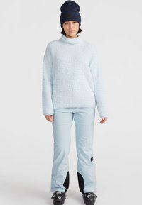 Lichtblauwe fleece pullover met een hoge nek, gecombineerd met lichtblauwe skibroeken met zwarte accenten en een marineblauwe beanie. Gladde textuur, relaxte pasvorm.