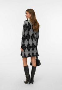 Donna in piedi, con un vestito argyle a maniche lunghe nero e grigio, stivali neri fino al ginocchio, che tiene in mano una piccola borsa nera.