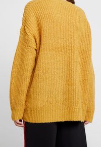 Hořčicově žlutý pletený svetr s volným střihem, žebrovanými manžetami a lemem. Texturovaný vzor s měkkou, hrubou vazbou. Zobrazen pohled zezadu.