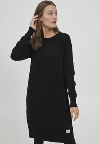 Schwarzes Strickkleid mit langen Ärmeln, Rundhalsausschnitt und lockerer Passform. Verfügt über ein kleines Logo-Patch auf der unteren rechten Seite.