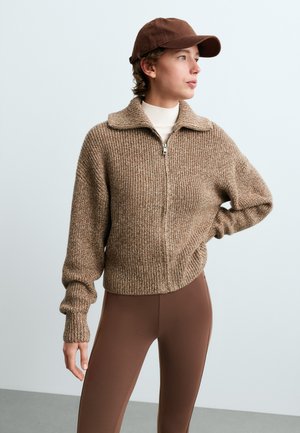 Personne portant un pull en maille marron zippé, une casquette marron et un pantalon marron ajusté, debout avec une main sur la hanche et regardant sur le côté.