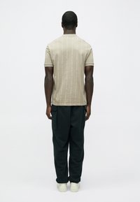 Camiseta beige de manga corta con patrón vertical sutil y cuello redondo, combinada con pantalones deportivos verde oscuro, vista desde atrás.