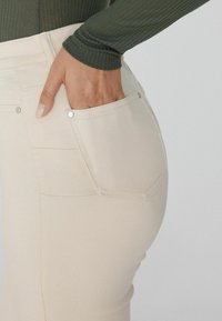 Jeans beige con forma ajustada, con un bolsillo trasero con herraje plateado. La tela tiene una textura suave, combinada con una blusa de canalé verde oscuro.