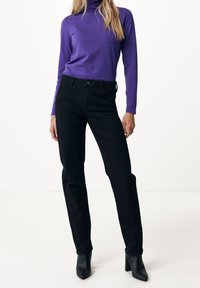 Svarta denimjeans med en slim-fit design, kombinerat med en långärmad lila polotröja. Modellen har på sig svarta klackade ankelstövlar.