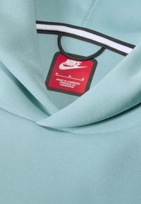 Šviesiai mėlynas Nike džemperis su lygiu paviršiumi, turi juodą raištelį ir dryžuotą akcentą prie kaklo. Matoma dydžio etiketė.