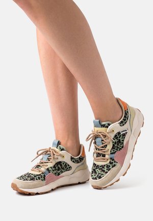 Füße in beige-pinken Turnschuhen mit grün-schwarzem Leopardenmuster und hellbraunen Schnürsenkeln vor weißem Hintergrund.