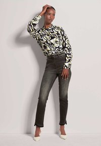 Langsleeve shirt met een zwart, geel en wit abstract patroon, gecombineerd met donkere, hooggesneden spijkerbroek en witte puntige pumps.