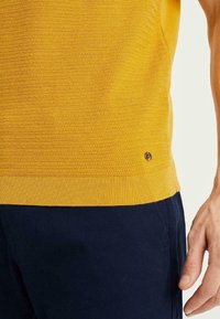 Yellow knitted sweater featuring a ribbed texture and a small logo detail at the hem, paired with navy blue pants.
