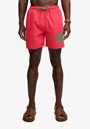 Hombre lleva pantalones cortos de baño de color coral brillante con cordón verde y texto verde parcial "SUP" en la pierna derecha, combinados con sandalias de cuero marrón.