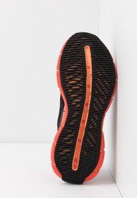 Semelle de chaussure de sport noire avec caoutchouc texturé pour l'adhérence, présentant une semelle extérieure orange vif et une bande orange contrastante au centre.