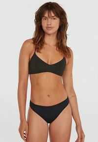 Donna con capelli castani uniformi e lunghi fino alla spalla, che indossa un bralette nero e slip bikini abbinati, in piedi contro uno sfondo chiaro e semplice.