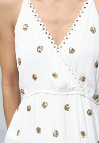 Robe blanche avec décolleté en V profond, bordure festonnée et embellissements en sequins dorés. Tissu texturé et taille cintrée.