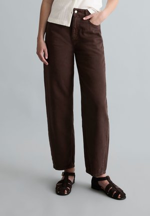 Personne portant un pantalon brun foncé à jambes droites et des sandales noires à bout fermé avec des brides à la cheville, main dans la poche.