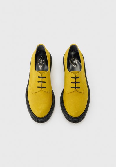 Paire de chaussures à lacets en daim jaune vif avec semelles noires, vue de dessus sur un fond gris clair.