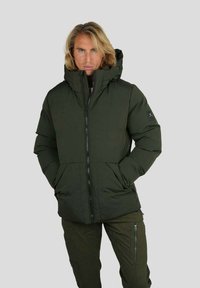 Personne blonde portant une veste rembourrée vert foncé avec col montant et un pantalon vert foncé assorti avec poches zippées, les mains dans les poches de la veste.