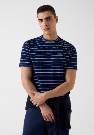 Młody mężczyzna o ciemnych włosach, ubrany w granatowo-białą pasiastą koszulkę GUESS oraz ciemny sweter związany wokół talii.