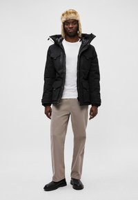 Chaqueta de invierno negra con cierres de cremallera y botones, dos bolsillos frontales, llevada sobre una camiseta blanca, combinada con pantalones beige claros y botas negras.