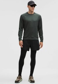 Zelená texturovaná košile s dlouhým rukávem, černé sportovní šortky, černé legíny, hnědé a zelené tenisky, čepice a sluneční brýle.