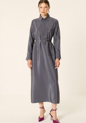 Femme portant une longue robe-chemise en satin gris avec une ceinture et un grand bouton, associée à des chaussures à talons hauts violettes avec lien à la cheville.