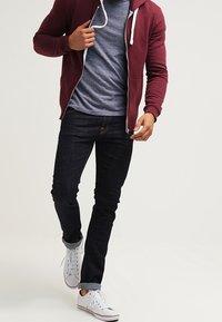 Mörkröd zip-up hoodie med vita snören, grå T-shirt under, mörka denimjeans och vita sneakers med uppvikta muddar.