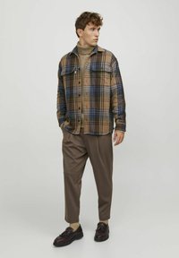 Giacca camicia a quadri marrone con dettagli blu, chiusura a bottoni e due tasche sul petto, indossata sopra un dolcevita beige e pantaloni marroni larghi.
