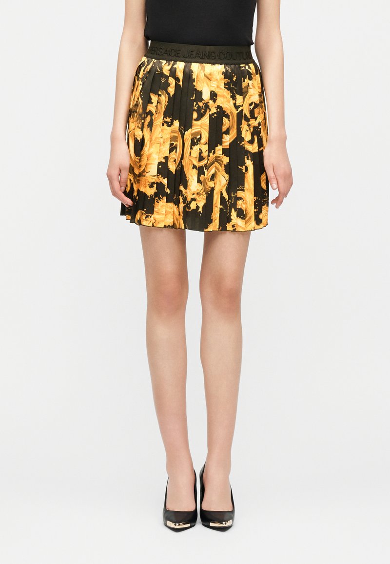 Γυναίκα που φοράει μαύρη πλισέ φούστα με φωτεινά κίτρινα μπαρόκ μοτίβα και μαύρες ψηλοτάκουνες γόβες με χρυσές λεπτομέρειες στα δάχτυλα.