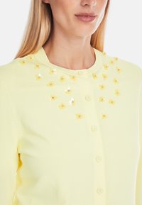 Light yellow cardigan with a round neckline and front buttons. Features floral appliqués with sequins for embellishment on the upper front.