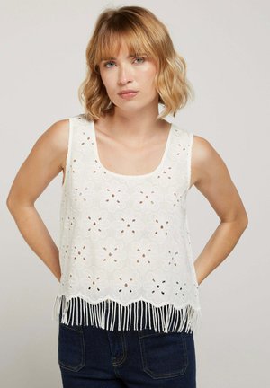 Femme aux cheveux blonds courts portant un haut sans manches en dentelle blanche avec des découpes florales et un ourlet à franges, associé à un jean en denim foncé.