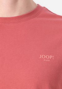 Rožinė medvilninė marškinėliai su apvalia iškirpte, kairėje krūtinės pusėje esantis "JOOP! JEANS" logotipas šviesiai rožinės spalvos. Lygus audinys, prigludusi forma.
