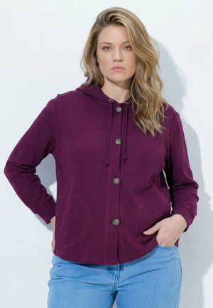 Kobieta z falowanymi blond włosami, ubrana w fioletową bluzę z kapturem na guziki i jasnoniebieskie jeansy, stojąca z jedną ręką w kieszeni na białym tle.