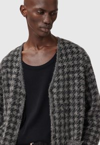 Grå og svart mønstret cardigan med strukturert strikk, med frontknapper og åpne kanter, lagdelt over en svart tank topp.