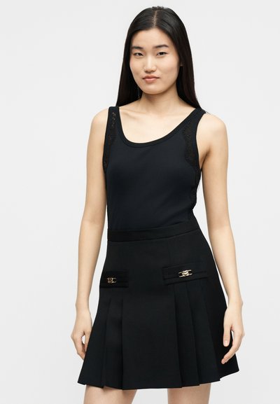 Γυναίκα με μακριά μαύρα μαλλιά φοράει ένα μαύρο αμάνικο τοπ με δαντελένιες λεπτομέρειες και μια μαύρη πλισέ φούστα με διακοσμητικές τσέπες.