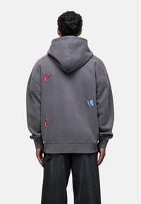 Sudadera gris de gran tamaño con capucha ajustable, que presenta bordados de mariposas coloridas en rosa y azul en la parte posterior.