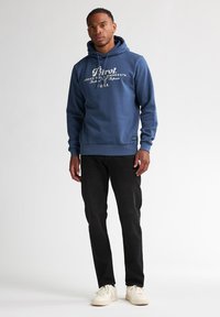 Felpa con cappuccio blu con stampa bianca, pantaloni di jeans neri e sneakers color crema. Stile casual, design semplice, e polsini e orlo a costine.