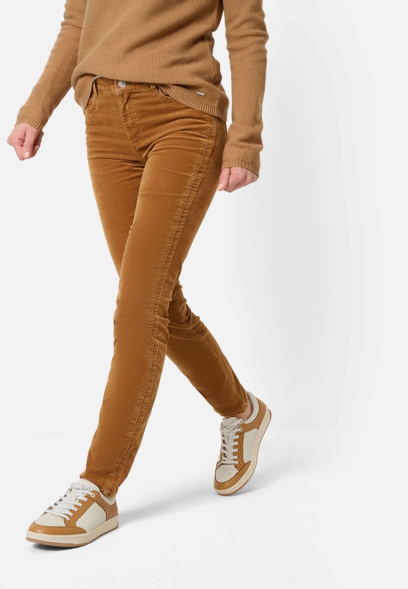 Personne marchant portant un pantalon en velours côtelé marron, un pull marron assorti et des baskets blanches et marron, les mains détendues le long du corps.