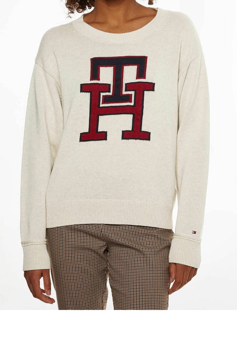 Kremowy sweter dziany z szerokim dekoltem, zdobiony dużym czerwono-granatowym logo. W połączeniu z brązowymi spodniami w kratę, podkreślającymi luźny krój.