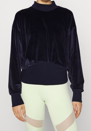 Person iført en mørk navy fløjlsbluse med lange ærmer med ribstrikkede manchetter og kant, kombineret med lyse leggings med gennemsigtige paneler.
