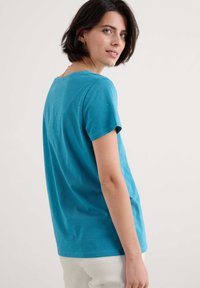 Türkisfarbenes Baumwoll-T-Shirt mit kurzen Ärmeln, lockerer Passform, gerundetem Ausschnitt und subtiler Textur. Der Rücken ist leicht länger als die Vorderseite.