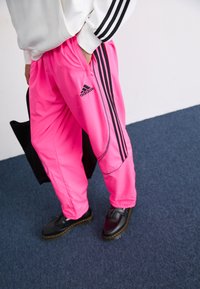 Bright pink track pants with black accents and three black stripes along the sides, paired with dark shoes. Lightweight, glossy material.
