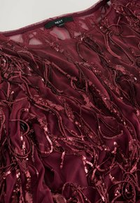 Burgunderfarbener transparenter Stoffoberteil mit langen, fließenden Strängen, die mit glänzenden Pailletten verziert sind und ein strukturiertes und geschichtetes Aussehen schaffen.