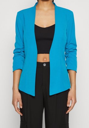 Person trägt ein leuchtend blaues Sakko über einem schwarzen, bauchfreien Top und hoch taillierte schwarze Hose mit Knopfverschluss.